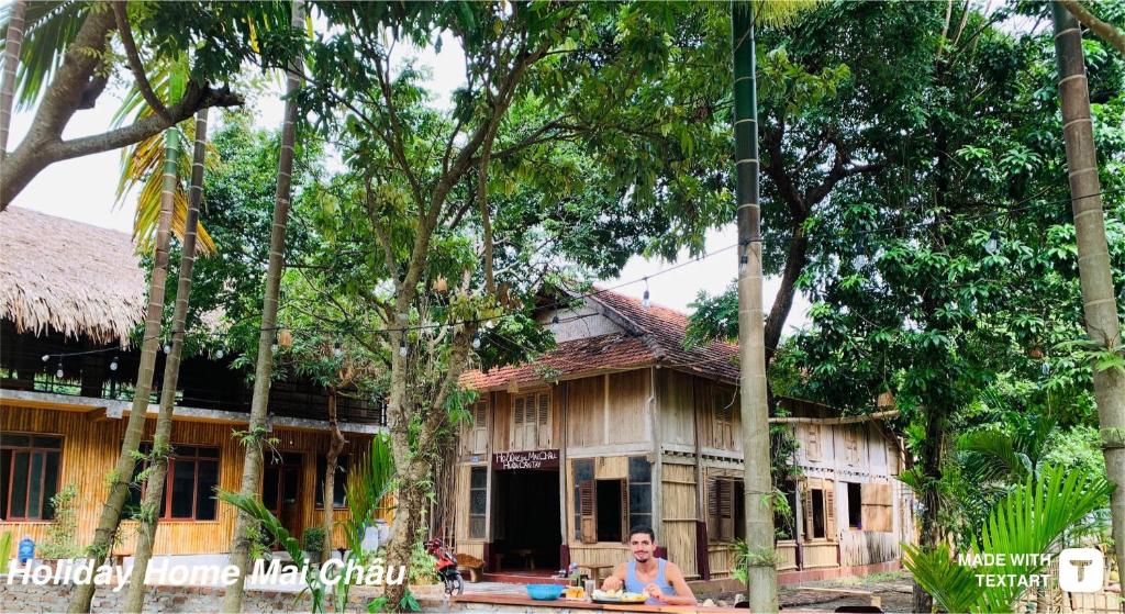 a man sitting in front of a house with trees at Mai Chau Home Holiday in Mai Châu