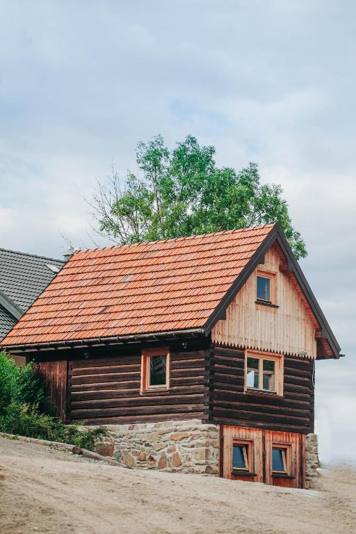 Rustic two-story wooden cabin with red tiled roof and stone foundation beside leafy tree.