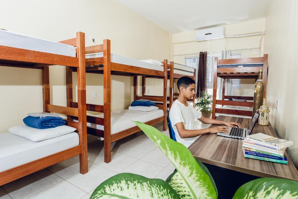 a man sitting at a desk with a laptop in a dorm room at Hotel Filhos da Promessa in Tucuruí
