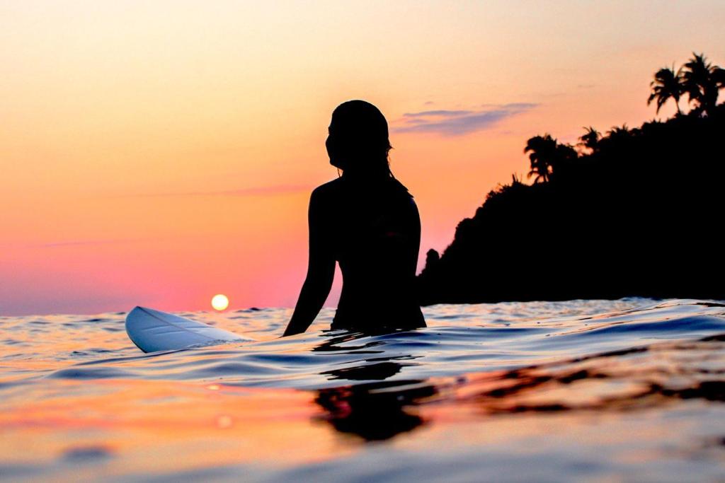uma mulher sentada numa prancha de surf no oceano ao pôr-do-sol em Secret Beach Hostel em Puerto Escondido