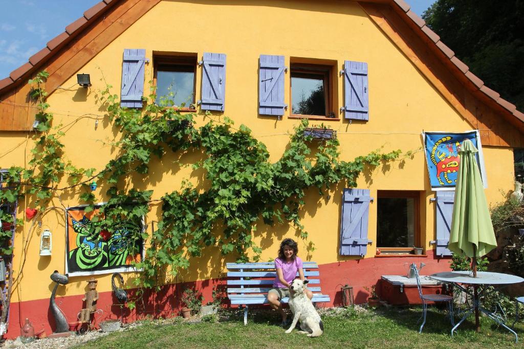 uma mulher sentada num banco em frente a uma casa com um cão em Abri de jardin aménagé, chambre Camping Glamour em Lalaye