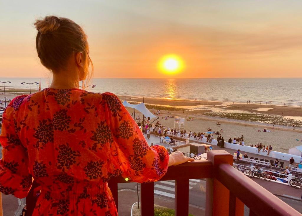 a woman standing on a balcony looking out at the beach at Hôtel Outre-Mer - Villa Le Couchant in Villers-sur-Mer