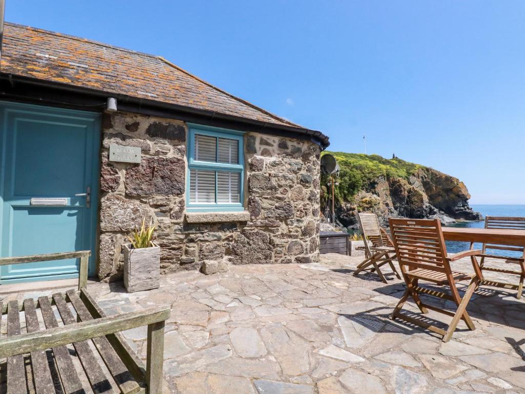 a stone cottage with chairs and a table and the ocean at Beach Cottage in Helston