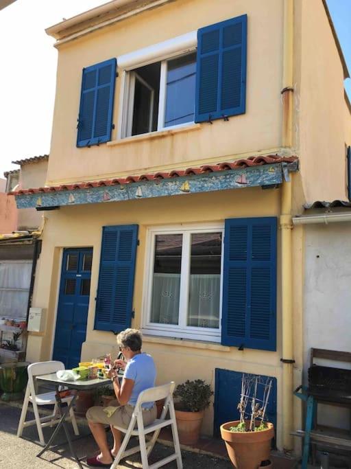 une personne assise à une table à l'extérieur d'une maison aux volets bleus dans l'établissement Cabanon aux Goudes Marseille calanques, à Marseille