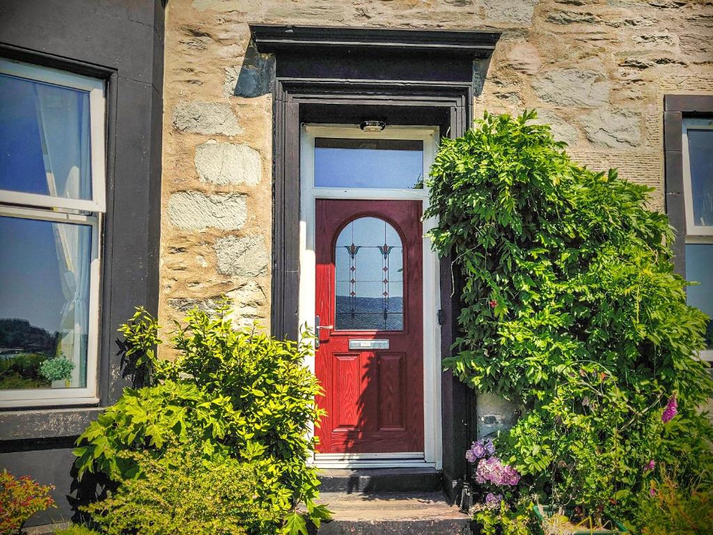 a red door on a stone house with two bushes at Firwood Apartment in Kilmun