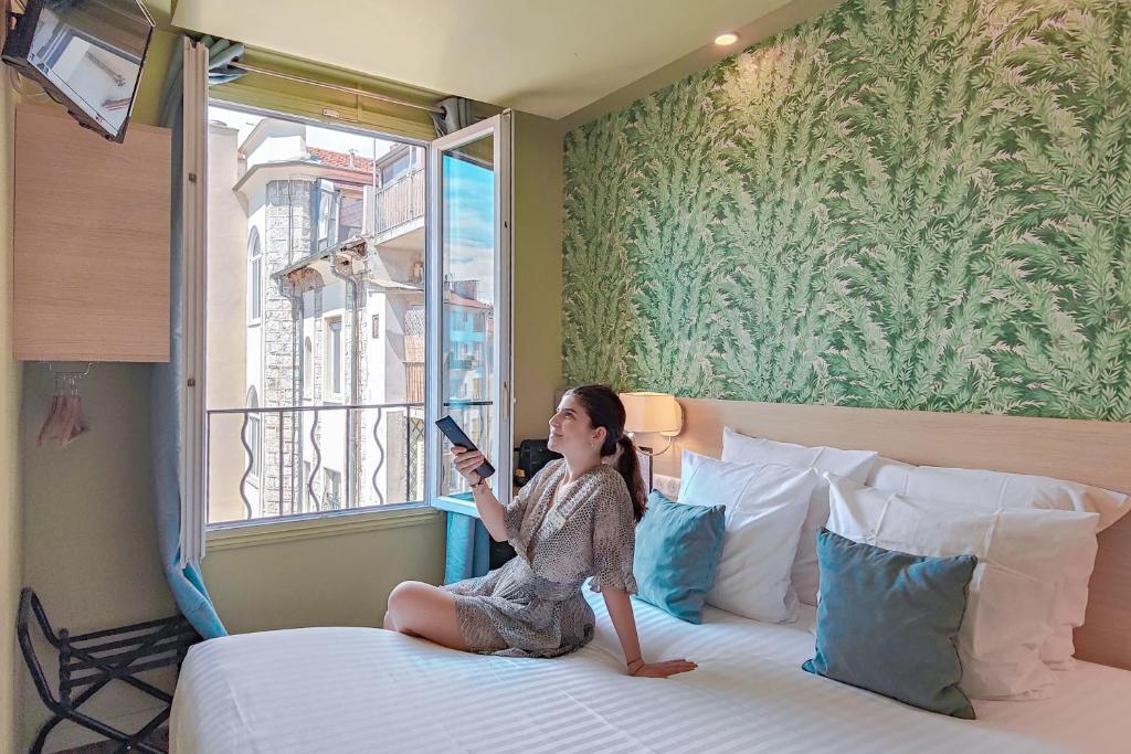 a little girl sitting on a bed looking out a window at Hôtel Du Centre, un hôtel AMMI in Nice