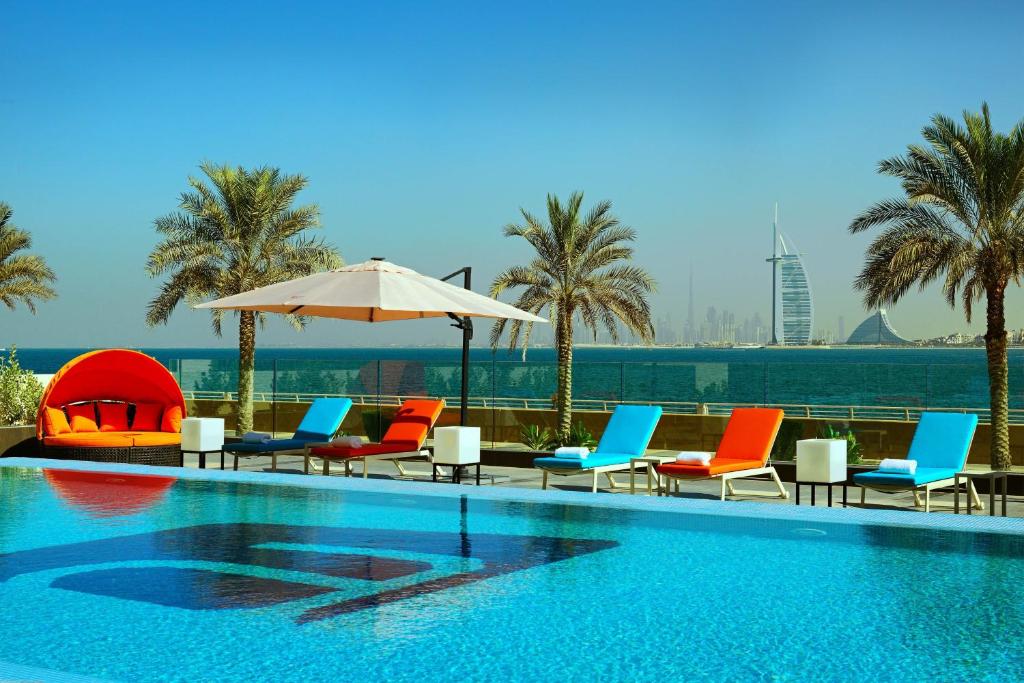 una piscina con sillas, sombrilla y palmeras en Aloft Palm Jumeirah en Dubái