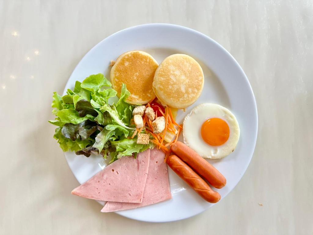A plated breakfast with a fried egg, sausages, ham, salad, vegetables and two round breads.