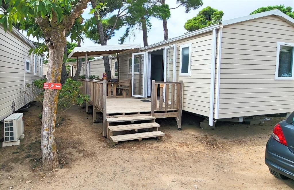 una casita blanca con un porche y un árbol en Mobil-home (Clim)- Camping Narbonne-Plage 4* - 019, en Narbonne-Plage