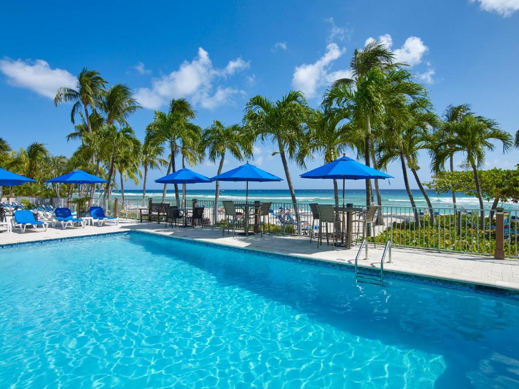 a swimming pool with palm trees and blue umbrellas at Coconut Court Beach Hotel in Bridgetown