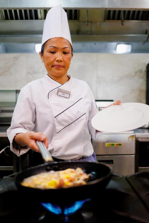 a woman in a kitchen preparing food in a pan at Tasia Maris Beach Hotel - Adults Only in Ayia Napa