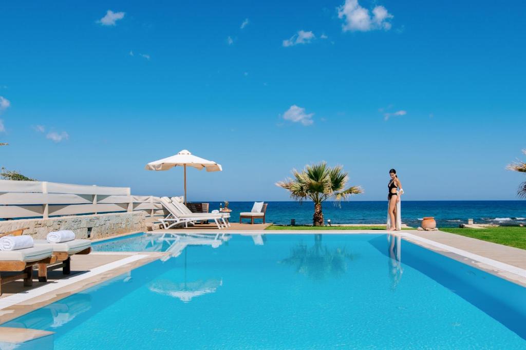 a woman standing next to a swimming pool next to the ocean at Hersonissos Beach Villa in Hersonissos