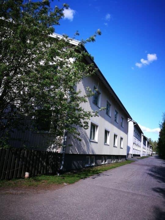 a white building with a fence next to a tree at -Hannula- Ilmastoitu persoonallinen ja rauhallinen kaksio kahdella autopaikalla kauppojen vieressä in Kuopio