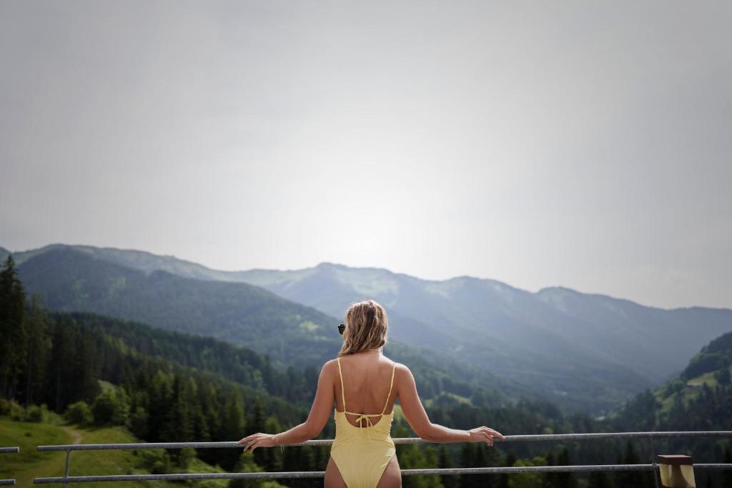 a woman in a yellow dress standing on a balcony looking at the mountains at Holzhotel Forsthofalm in Leogang