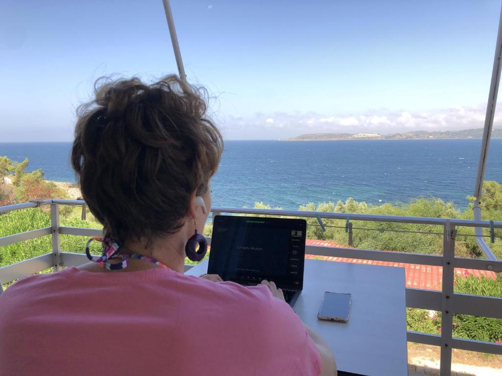 a woman sitting at a table with a laptop looking at the ocean at Galini Studios and Apartments in Lassi