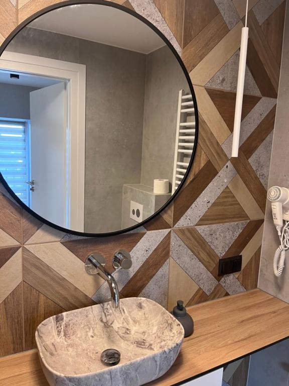 Stone sink beneath large round mirror with geometric wooden tile accent and modern pendant light.