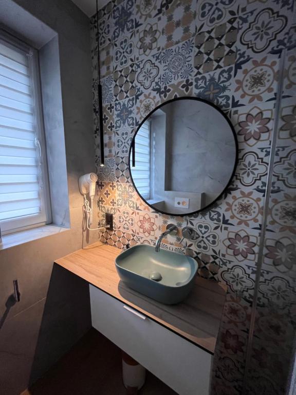 Bathroom sink and teal basin beneath round mirror, patterned tile wall and mounted hairdryer.