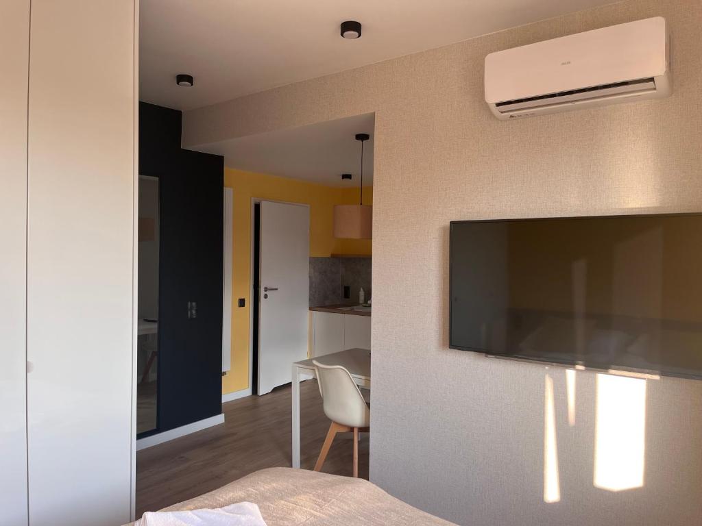 Room view showing wall-mounted TV, air conditioner, small dining table and kitchenette doorway, soft natural light.