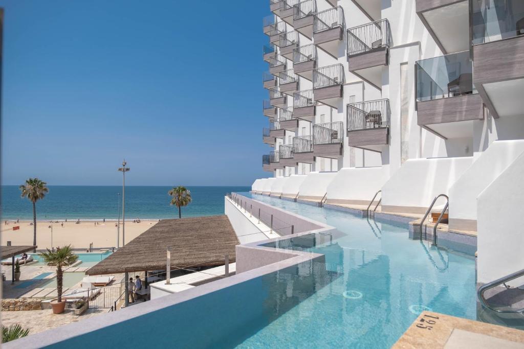 カディスにあるCádiz Bahía by QHotelsのスイミングプールとビーチのあるホテルの外観を望めます。