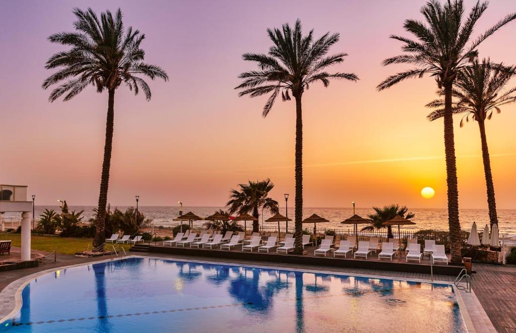 uma piscina com palmeiras e o oceano ao pôr do sol em Sea Life Nahariya BY Jacob Hotels em Nahariya