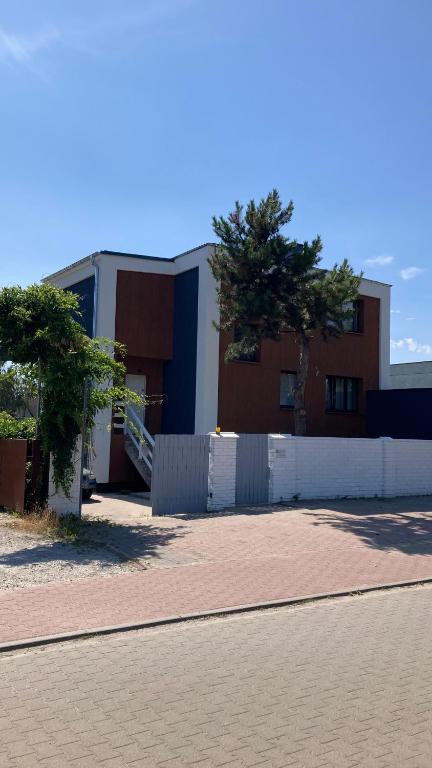 Modern two-story house behind white fence, exterior stairs, and small tree shadowed on sidewalk.
