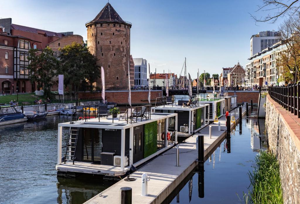 a row of boats docked in a river with a tower at Domy Na Wodzie - La Mare Motlava Houseboats with Sauna by Downtown Apartments in Gdańsk