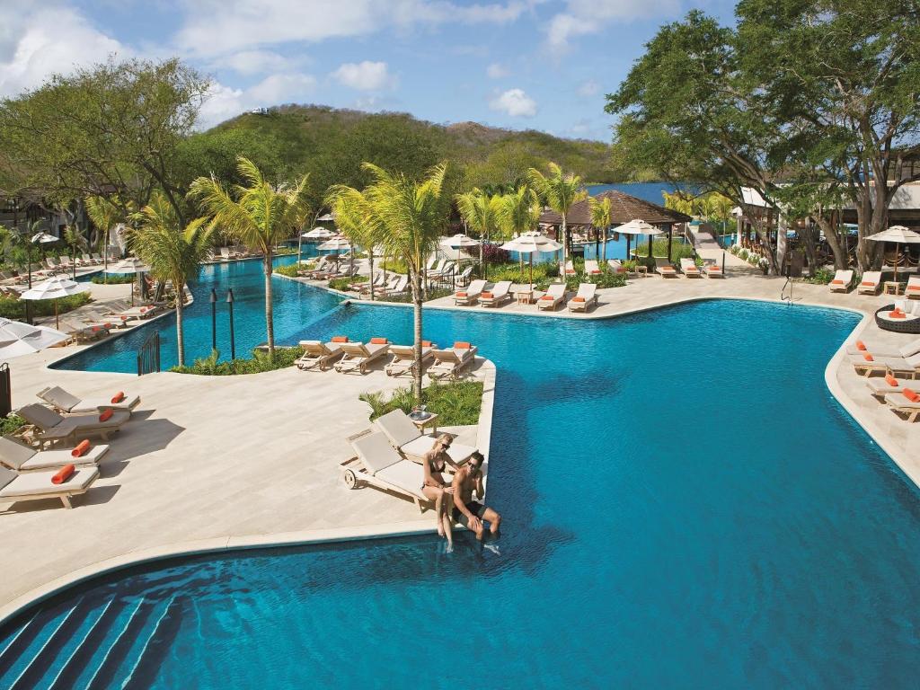 Interconnected freeform pools with palm-fringed sundeck, orange-cushioned loungers and umbrellas by a forested hillside at JW Marriott Costa Elena Resort & Spa, All-Inclusive in El Jobo
