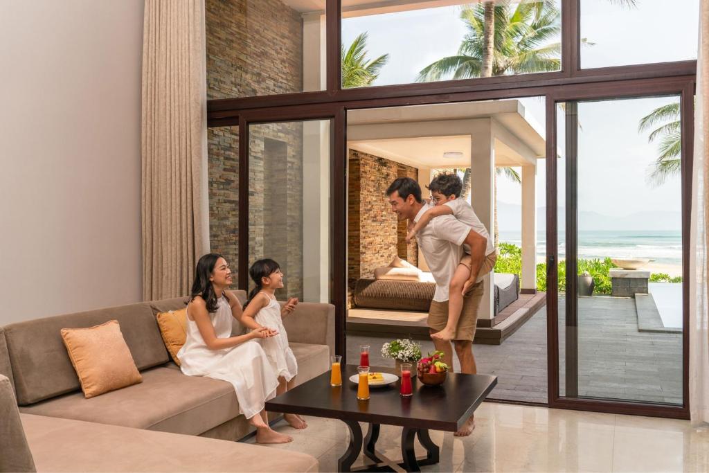 A family gathers in a living room opening onto a covered outdoor daybed and the sea.