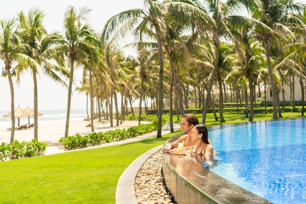 Two guests relax at the edge of an infinity pool amid palms beside the beach.