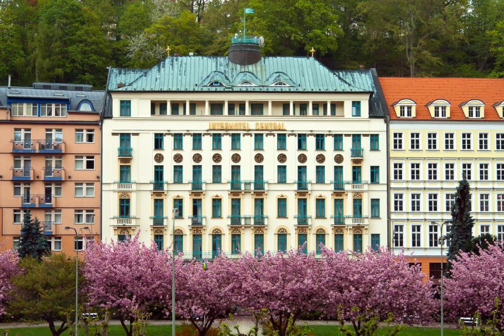 a large white building with a green roof and purple trees at Interhotel Central in Karlovy Vary