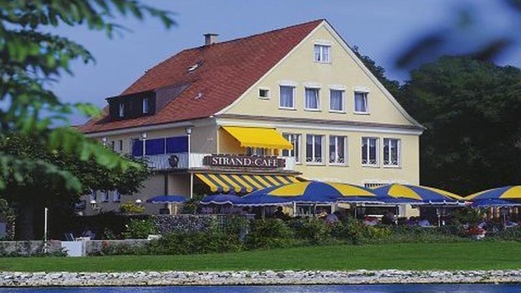un grand bâtiment avec des parasols bleus et jaunes devant lui dans l'établissement Hotel Strand-Café mit Gästehaus Charlotte, à Langenargen