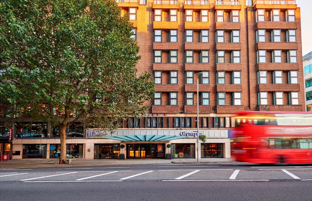 un autobús rojo conduciendo por una calle frente a un edificio en Hilton London Olympia, en Londres