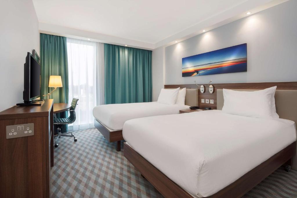 a hotel room with two beds and a desk at Hampton By Hilton London Stansted Airport in Stansted Mountfitchet