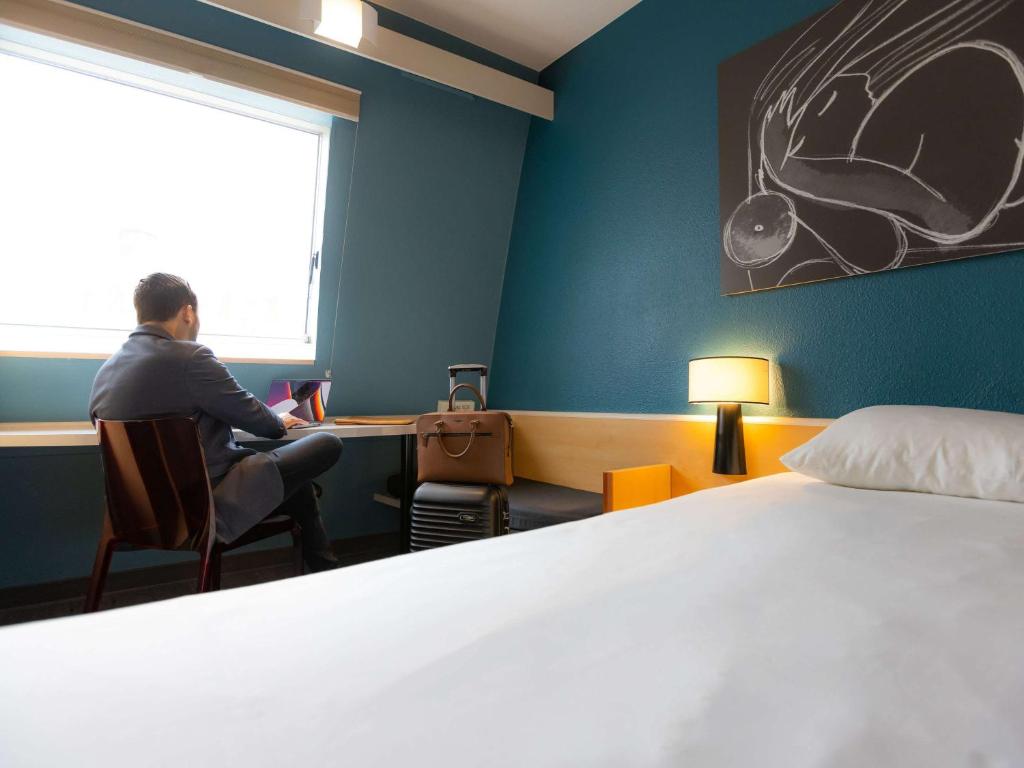 un homme assis à un bureau dans une pièce avec un lit dans l'établissement ibis Besançon Centre la City, à Besançon