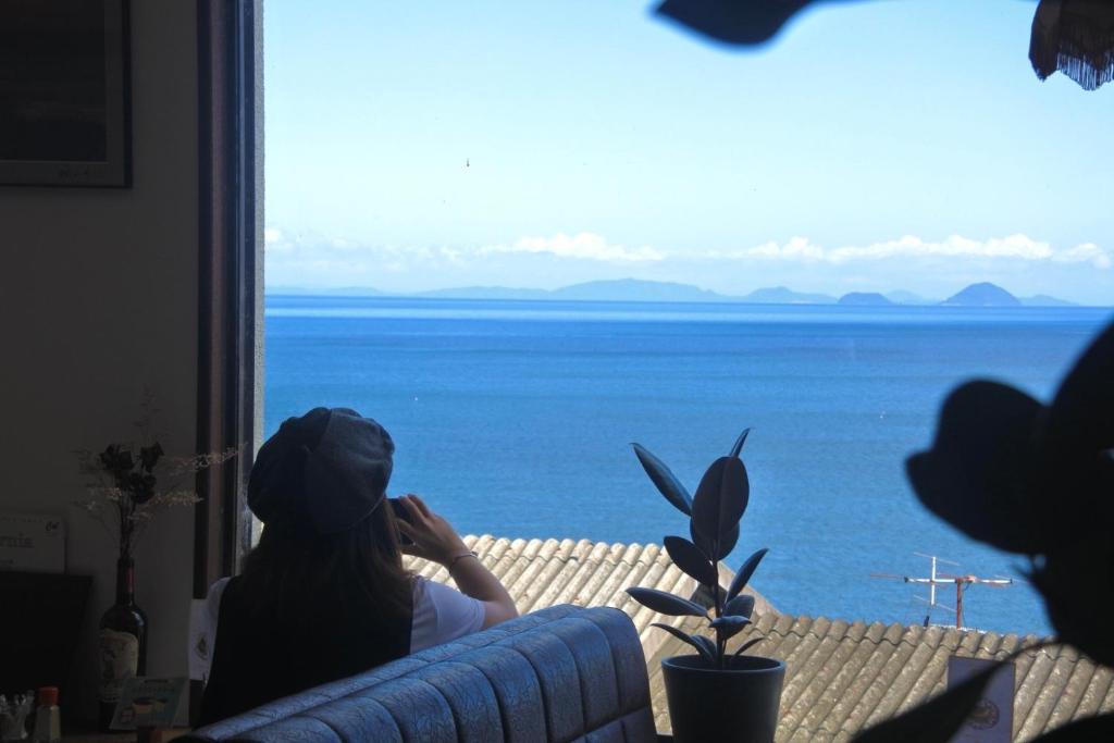 a woman sitting on a couch looking out a window at the ocean at 海に恋する泊まれる喫茶店 ポパイ in Iyo