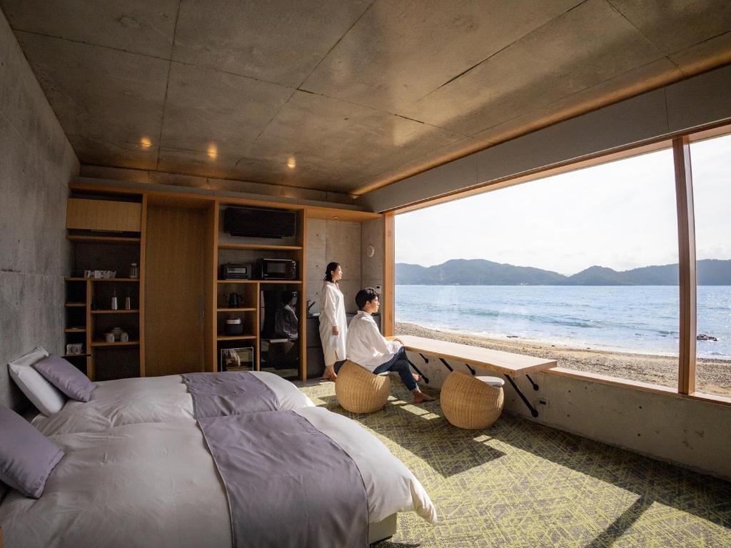 a man sitting in a bedroom with a view of the ocean at Denpaku The Beachfront MIJORA in Amami