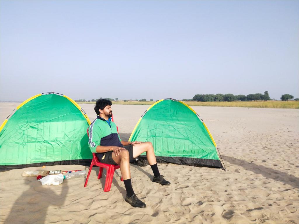 un hombre sentado en una silla junto a dos tiendas de campaña en la playa en Jhoomke camping and water sports adventure, en Auraiya