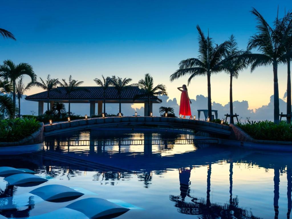 Una donna con un vestito rosso che attraversa un ponte sopra una piscina di Oriental Hotel Okinawa Resort & Spa a Nago