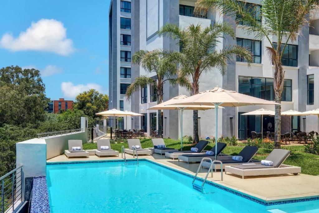 a swimming pool with chairs and umbrellas next to a building at Stay Regency Menlyn in Pretoria