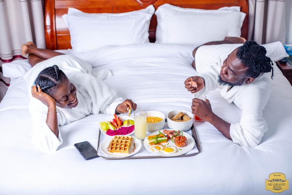 Ein Mann und eine Frau im Bett mit einem Tablett Essen in der Unterkunft MJ Grand Hotel in East Legon