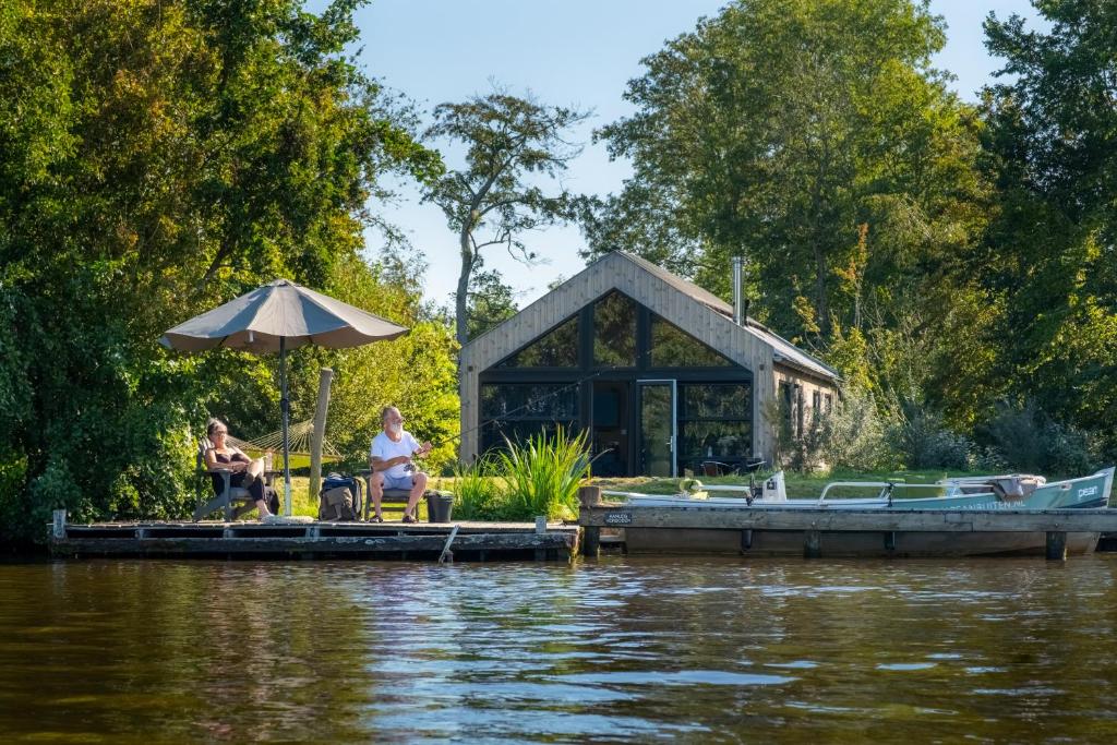 two people sitting on a dock with a house on the water at Pean-buiten Waterlodges in Nes