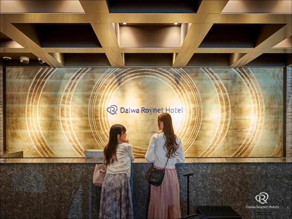 zwei Frauen stehen vor einer Wand mit einem Schild in der Unterkunft Daiwa Roynet Hotel Kyoto Shijo Karasuma in Kyoto