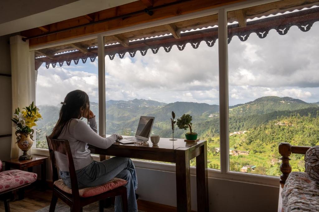 une femme assise à une table avec un ordinateur portable devant une fenêtre dans l'établissement SaffronStays The Clairmont Cottage,Mukteshwar, à Mukteswar