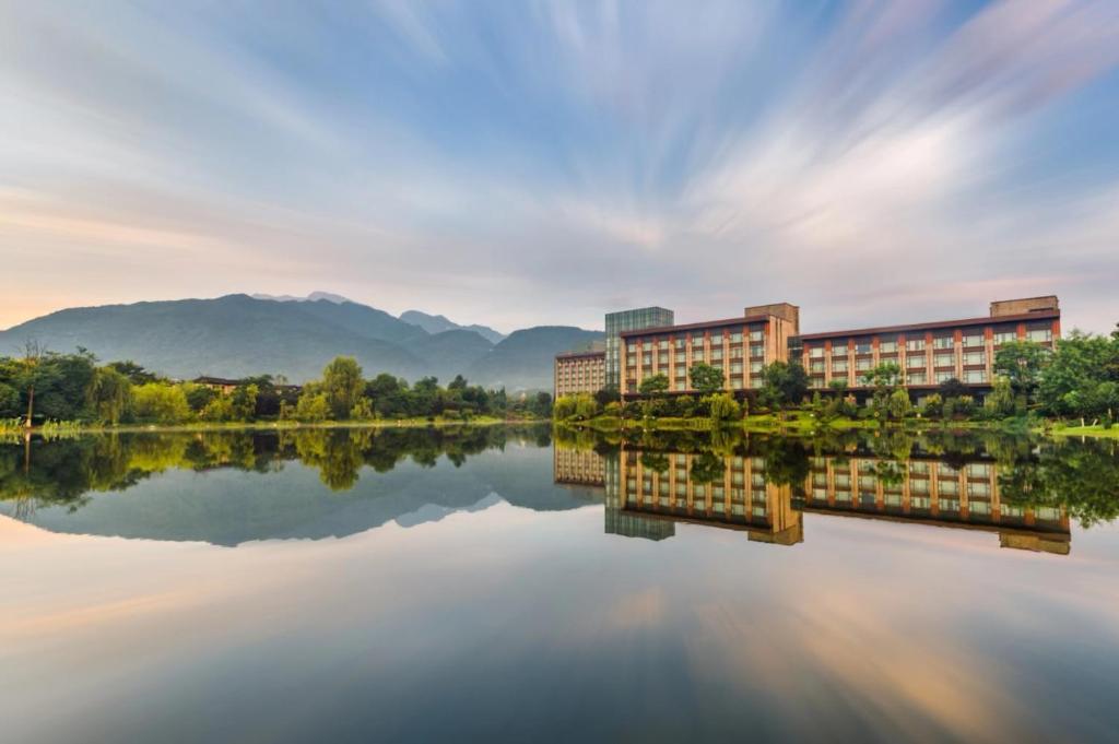 a building next to a lake with mountains in the background at Le Meridien Emei Mountain Resort in Emeishan