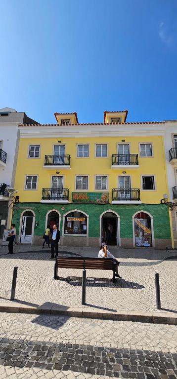 eine Person, die auf einer Bank vor einem gelben Gebäude sitzt in der Unterkunft BBL APARTAMENTOS - B in Nazaré