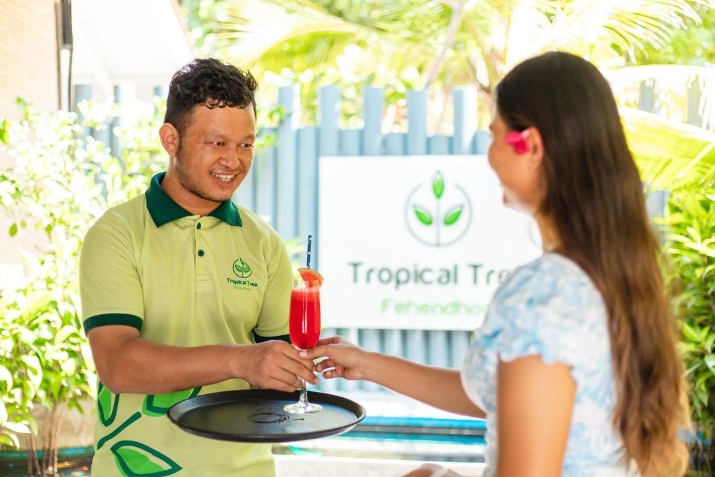 Un uomo che tiene un vassoio con una donna che regge un drink di Tropical Tree a Fehendhoo