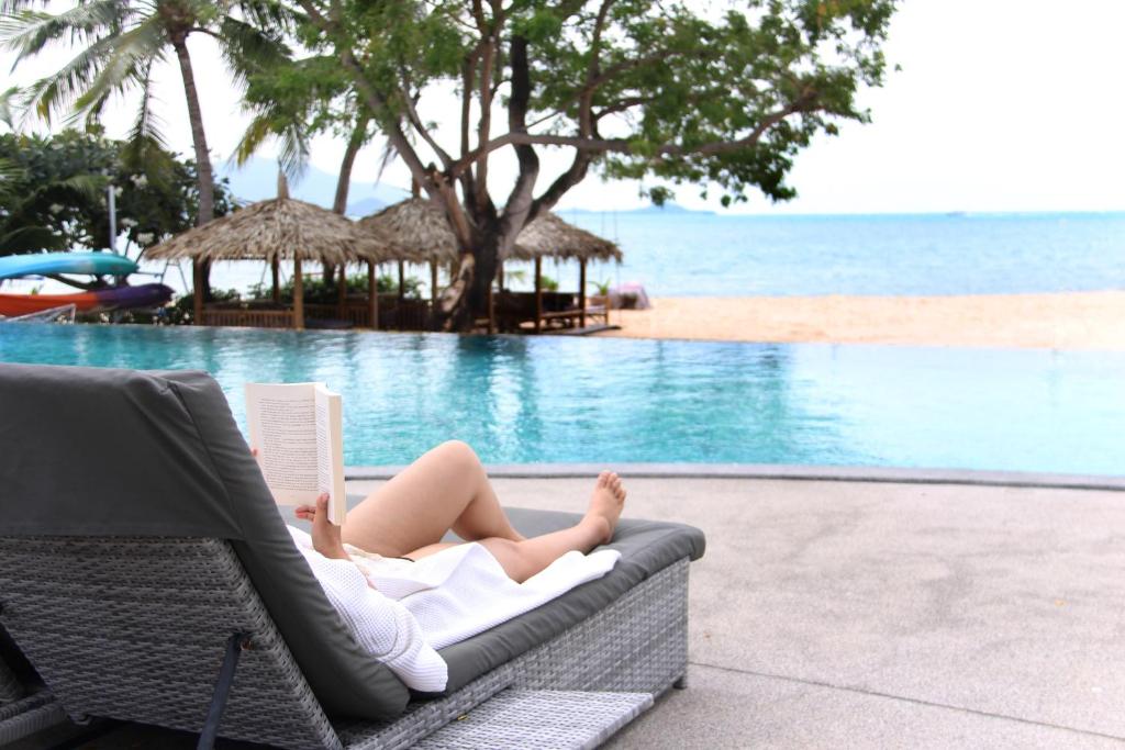 a woman laying on a chair reading a book next to a pool at Deva Beach Resort Samui in Choeng Mon Beach