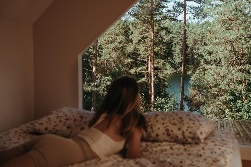 Una mujer tendida en una cama mirando por la ventana en Polana Gawrycha, domki nad jeziorem z widokiem en Suwałki