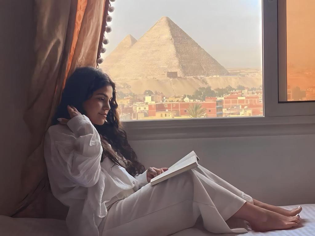 una chica sentada en una cama leyendo un libro frente a la pirámide en Crowne Pyramids view inn en El Cairo