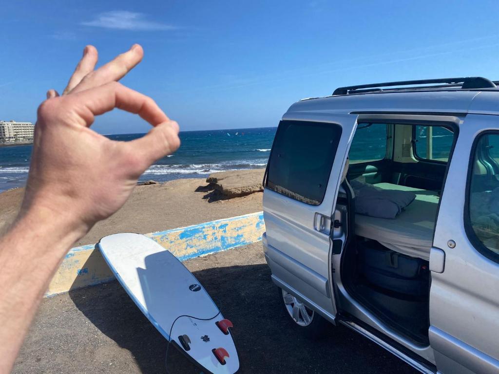 a person making a peace sign with a surfboard next to a van at CamperTF - old but lovable mini caravans in Tenerife in El Médano
